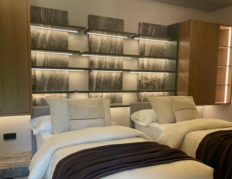 Dormitorio con dos camas y, detrás, estantes iluminados sobre un fondo de revestimiento efecto piedra