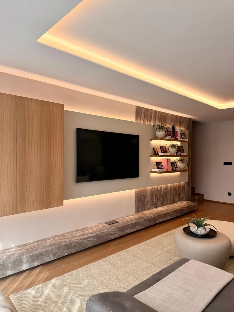 Salón con la pared de la televisión revestida en efecto piedra y madera, estantes iluminados y un techo con luz indirecta