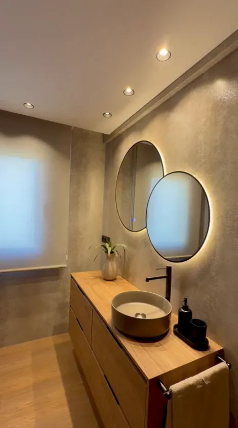 Vídeo vertical de un recorrido por un baño con paredes de revestimiento continuo, un mueble de madera, espejos redondos y una mampara de ducha